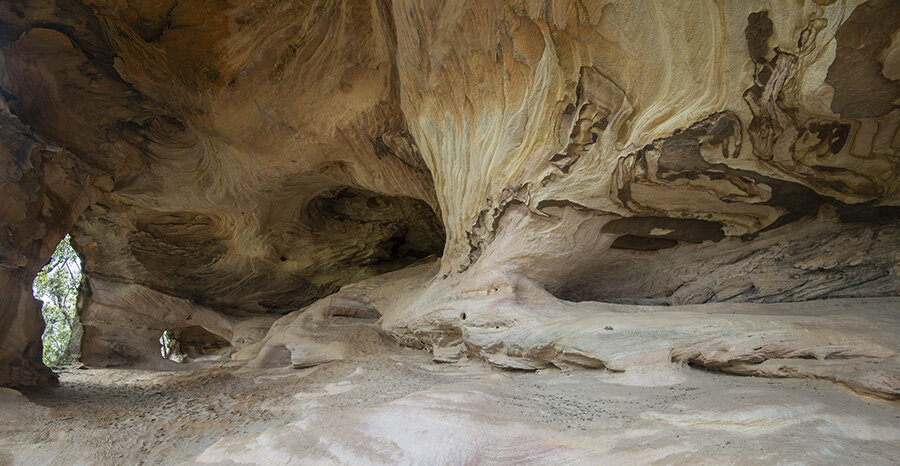The Grand Cathedral: interior of a large sandstone cave with curved ochre walls, an arching ceiling, and a sunlit opening to greenery.