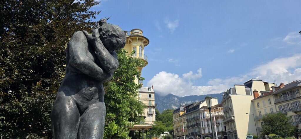 A dark bronze sculpture of two curled figures in a park at Aix-les-Bains, with a yellow belle époque villa and mountains behind