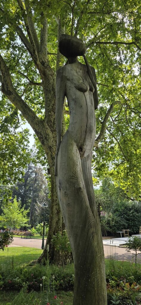 Tall wooden sculpture of a slender female figure carved from a single trunk, standing under a plane tree in the public gardens at Aix-les-Bains