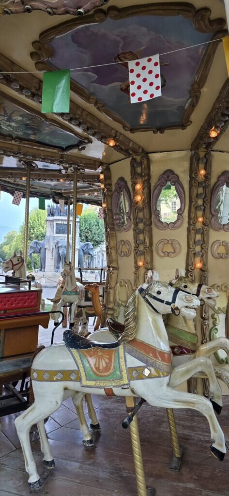 A closed vintage carousel in Chambéry with painted wooden horses in mid gallop, and the Fontaine des Éléphants visible in the gap behind them