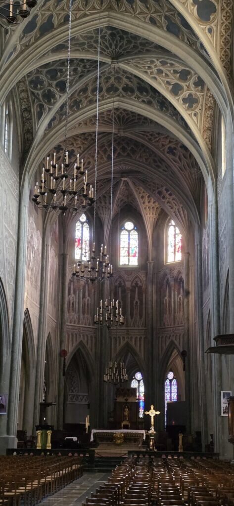 The tall nave of Chambéry cathedral with rows of empty wooden chairs, hanging candle chandeliers and stained glass windows at the far end