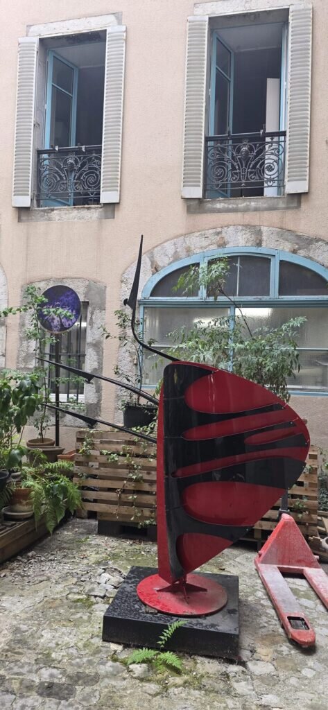 A large red and black curved steel sculpture standing on a stone plinth in a cobbled Chambéry courtyard, with potted ferns, wooden pallets and shuttered windows behind