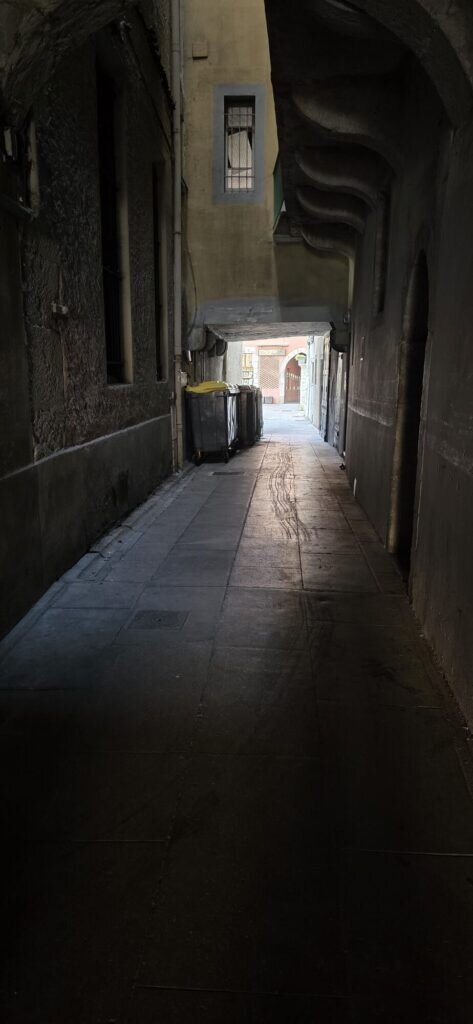 A dark covered stone passage running under buildings in Chambéry, with bins along one wall and daylight at the far end