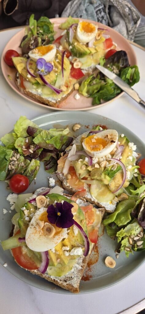 Two plates of open sandwiches topped with soft boiled egg, avocado, red onion, hazelnuts and an edible purple violet, with salad on the side