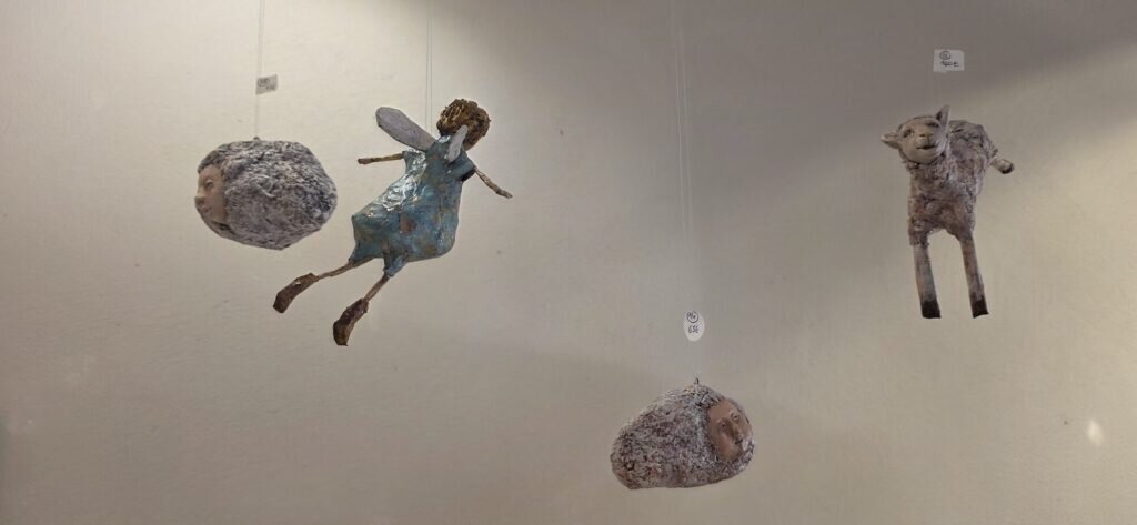 Ceramic sculptures suspended on wires against a white gallery wall, including a winged girl in a blue dress, a lamb, and two sleeping heads on clay clouds