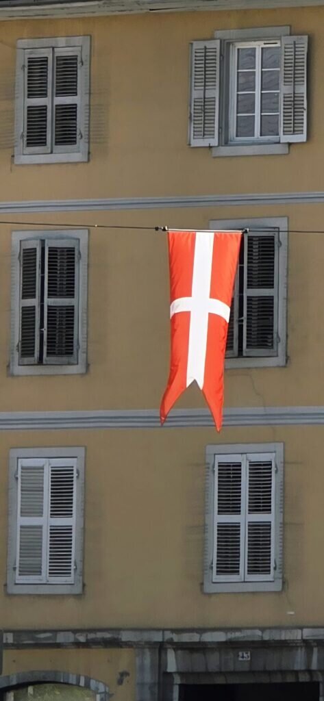 A red banner with a white cross, the flag of Savoy, hanging from a wire in front of a yellow building with grey shutters