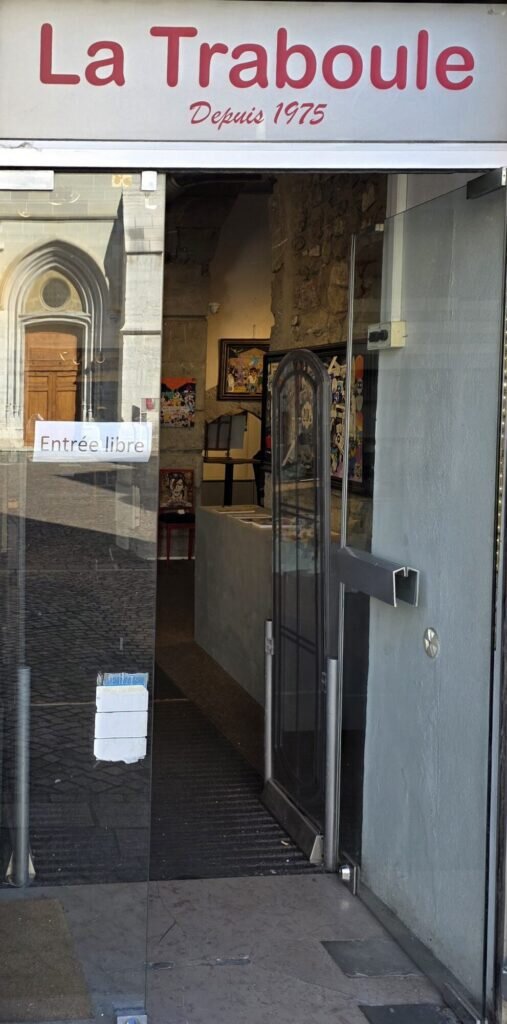 A glass shopfront door in Chambéry under a sign reading La Traboule depuis 1975, with framed artwork hanging inside a stone passage and a gothic church doorway reflected in the glass
