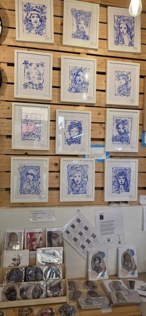 Twelve framed blue ink portraits of faces surrounded by flowers on a slatted wooden wall, with a rack of printed cards, coasters and notebooks below