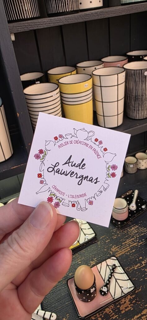 A hand holding the printed card of ceramicist Aude Lauvergnas in front of shelves of striped and checked faience beakers