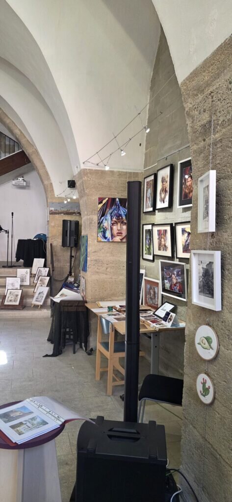 Inside the vaulted Grenier Public exhibition space, framed paintings and drawings on stone walls with track lighting and embroidery hoops