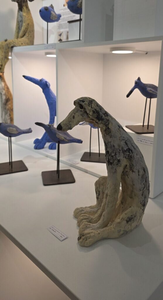 A pale ceramic sitting hound with a long nose on a gallery shelf, surrounded by small blue ceramic birds and dogs on metal stands