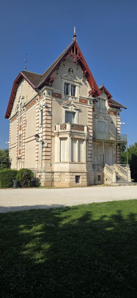 A nineteenth century villa with a steep red trimmed roof and shuttered windows standing in the gardens of Parc Gautier, L'Isle-sur-la-Sorgue