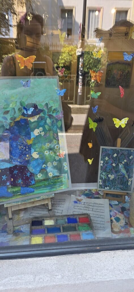 A gallery window in L'Isle-sur-la-Sorgue hung with cut paper butterflies on wires, painted panels of blue and green gardens inside, and a compartmented wooden tray of colour blocks on the sill
