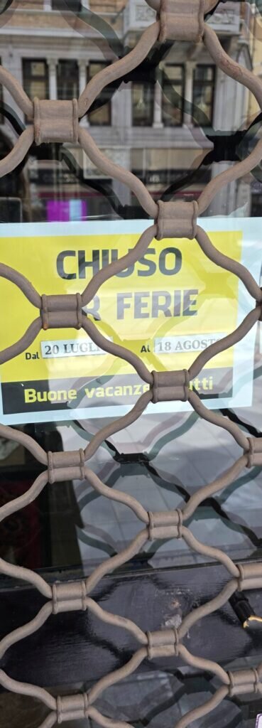 A yellow closed for holidays sign reading chiuso per ferie behind a decorative metal grille on a Turin shop door