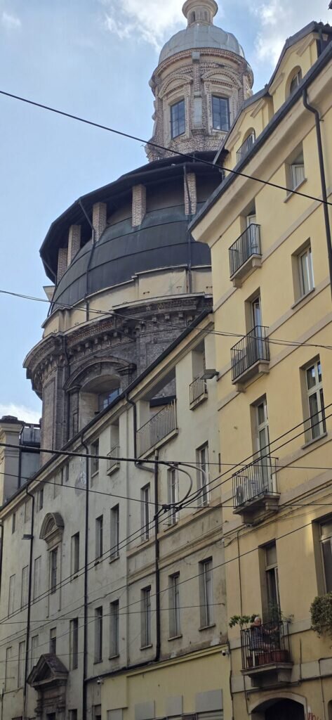 A great dark church dome rising above ochre buildings at the end of a straight Turin street, tram wires crossing the view