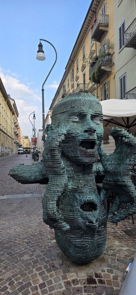 A large modern bronze sculpture of a fragmented, pixelated human head with an open mouth and raised hands on a Turin street