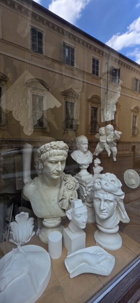 A Turin antique shop window full of white plaster busts, classical heads and cherubs, with the street reflected in the glass