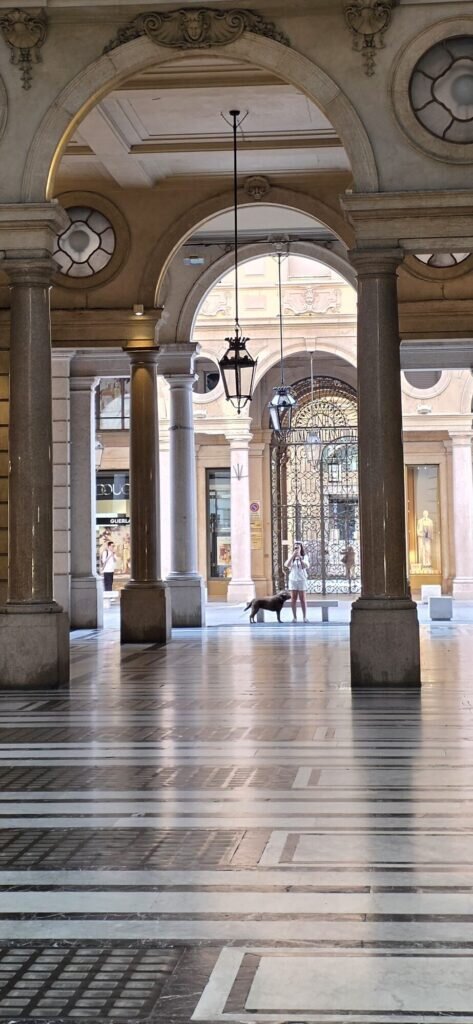 A Turin portico arcade with a patterned black and white marble floor running into the light, a distant figure with a small dog under the arch