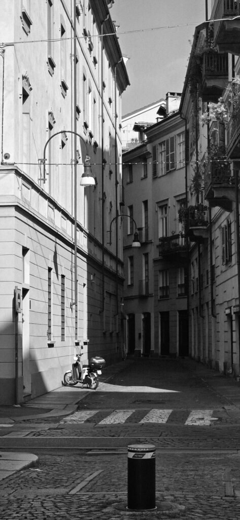 Black and white photograph of an empty narrow lane in Turin's Roman quarter, shutters closed, a single scooter parked against the kerb