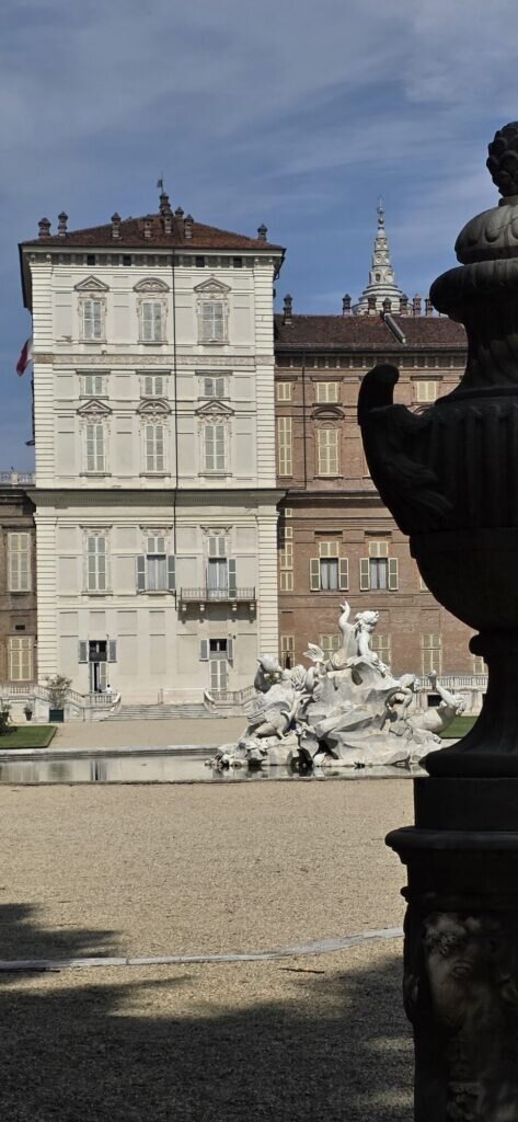 The white facade of the Royal Palace in Turin seen from the Royal Gardens with the marble Tritons fountain in front and a large stone urn in the foreground