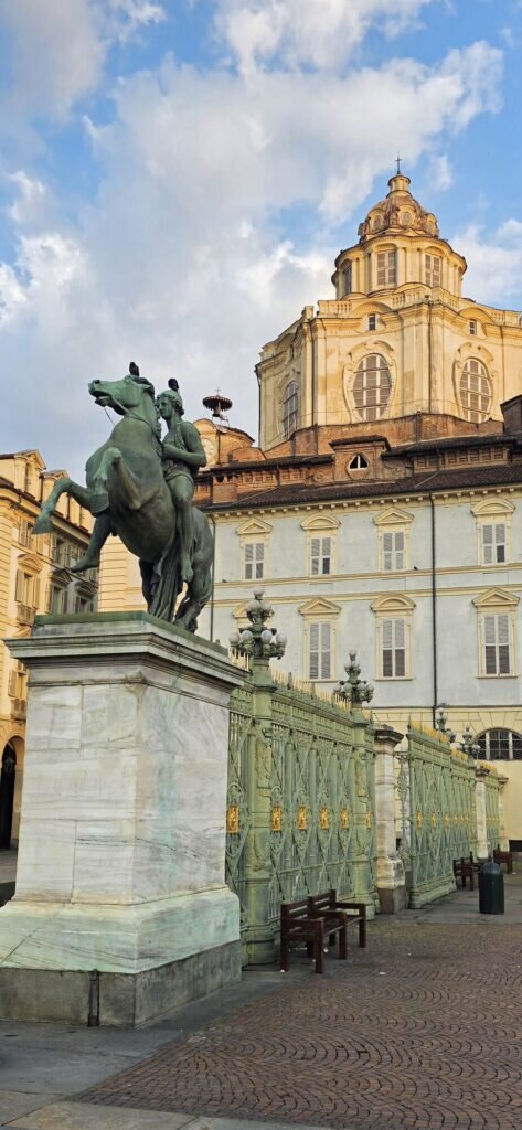 A rearing bronze horseman at the gates of Turin's Royal Palace with the gold Baroque dome of San Lorenzo behind in early morning light