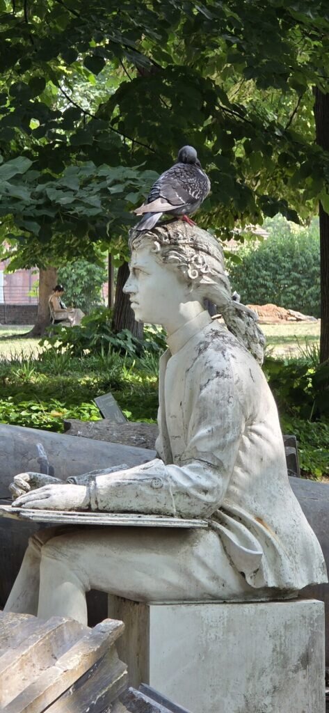 A weathered white marble statue seated with a board on its lap in a Turin garden, a pigeon standing on top of its head
