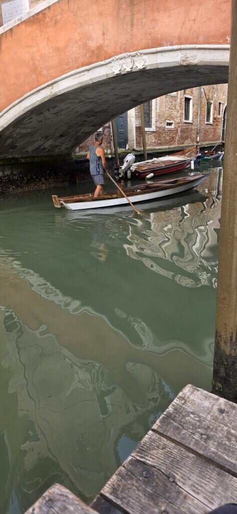 A man rowing a small wooden boat under a low stone bridge on a Venetian canal, standing and using a single oar