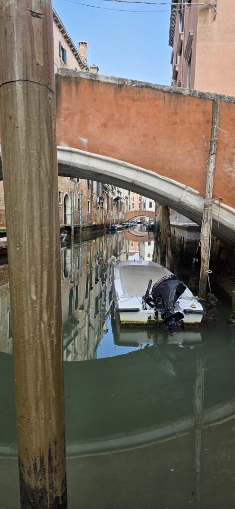 A Venetian canal seen through an arched bridge with striped mooring poles and a moored white motorboat, doubled in reflection
