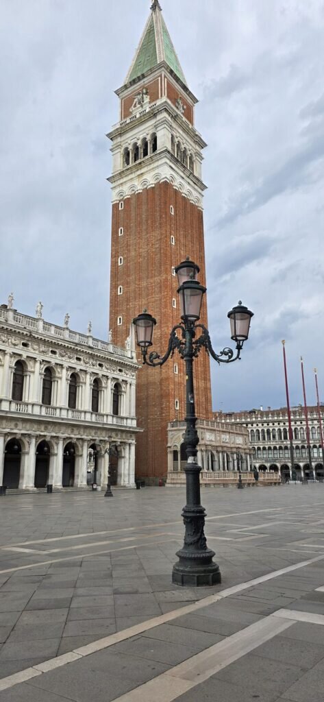 The campanile bell tower of St Mark's rising over a nearly empty Piazza San Marco at dawn, with an ornate iron lamp post in front