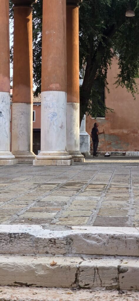 A quiet Venetian campo with orange colonnade columns and a distant figure sweeping the stone paving
