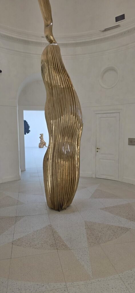 A glossy gold ribbon-like abstract sculpture inside a white pavilion interior