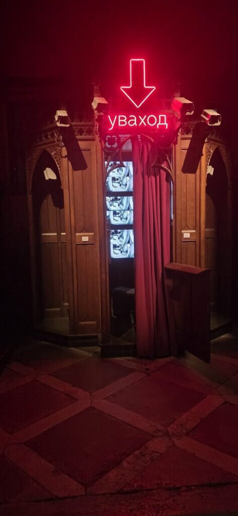 A dark red exhibition space with glowing neon Cyrillic signage above small screens and old wooden pews
