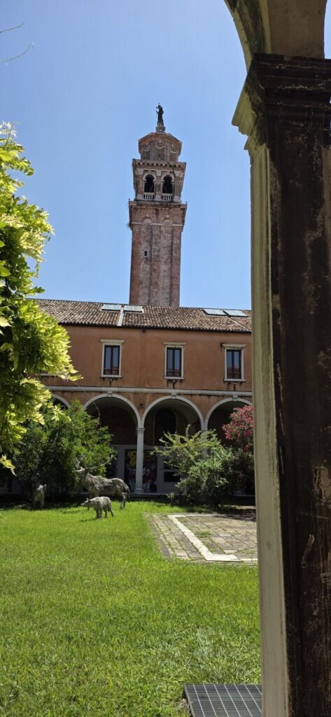A leaning brick bell tower rising above a cloister garden with pale animal sculptures on the lawn
