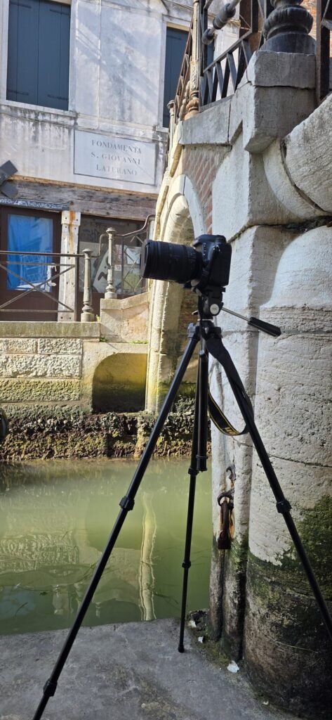 A camera on a tripod set up beside a canal near the Ospedaletto church in Castello, Venice, with a street sign reading Fondamenta San Giovanni Laterano