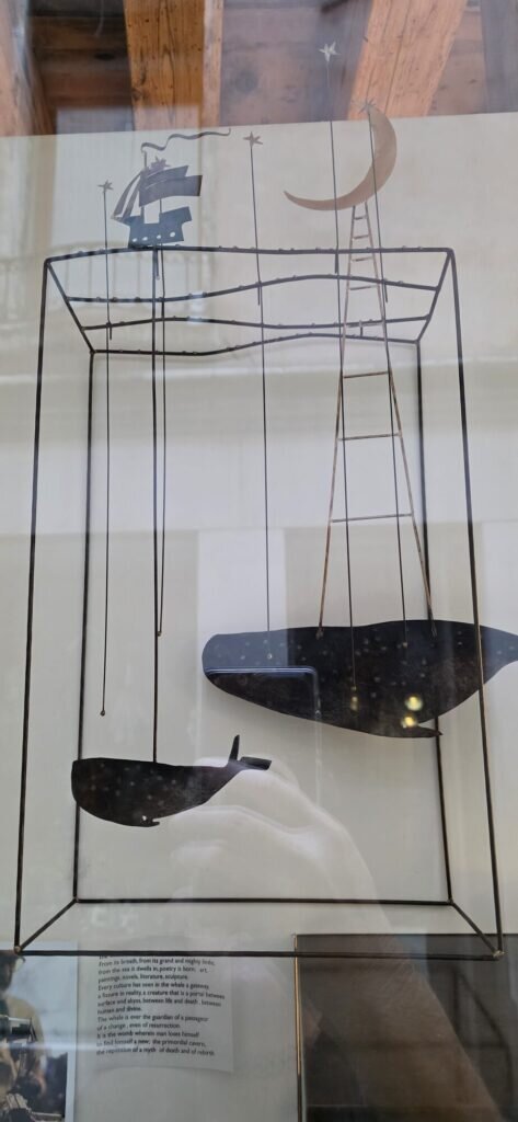 A wire and cut-metal sculpture behind glass showing two black whale shapes, a small ship and a crescent moon on a ladder