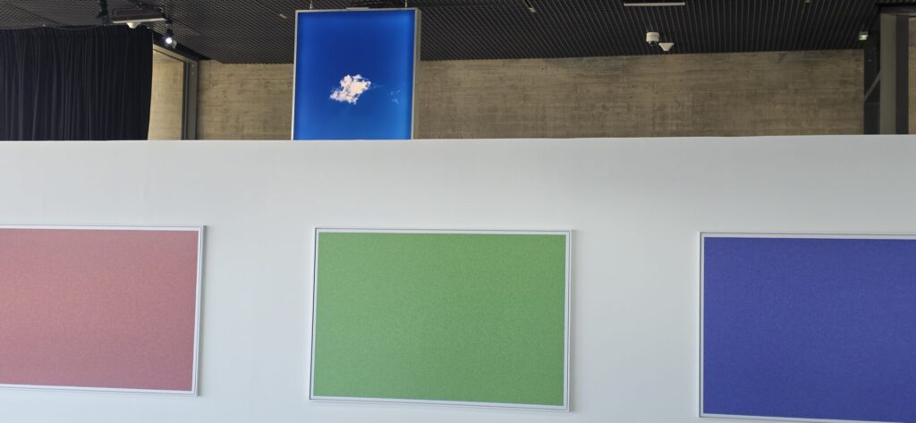 A white gallery wall hung with three large framed monochrome panels in dusty pink, green and blue, with a photograph of a single small cloud in a deep blue sky mounted on the concrete wall above
