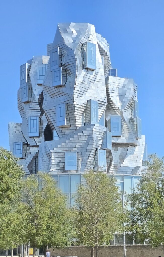 The LUMA Arles tower by Frank Gehry, a twisting stack of stainless steel blocks above a glass base, seen over young trees