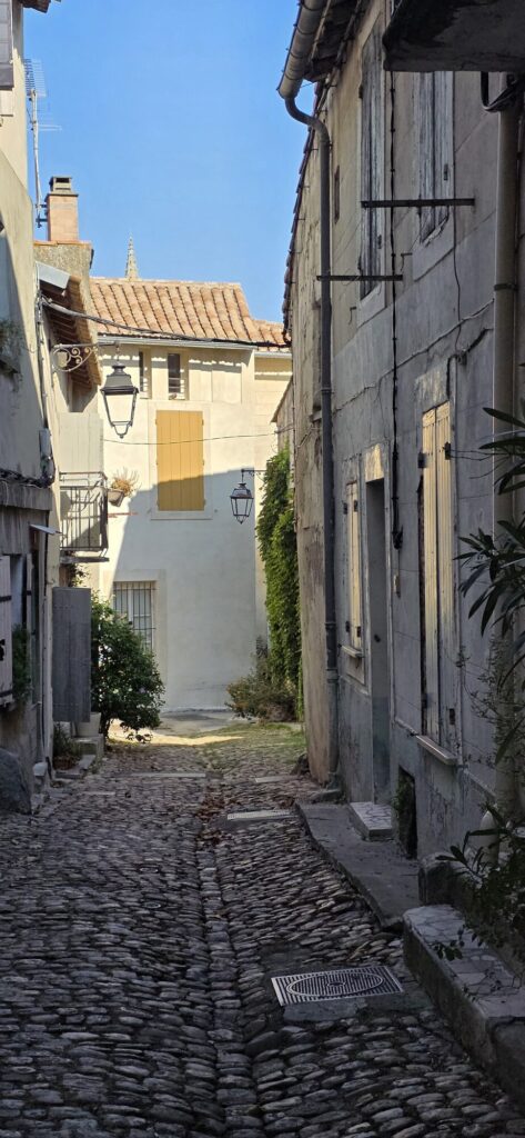 An empty cobbled lane in Arles in early morning light, with shuttered stone houses, a yellow shutter and two iron street lanterns