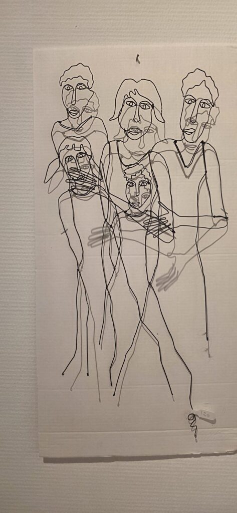 A wire sculpture of four figures hung flat on a white gallery wall, casting a soft grey shadow that doubles every line, with a small price tag at the corner