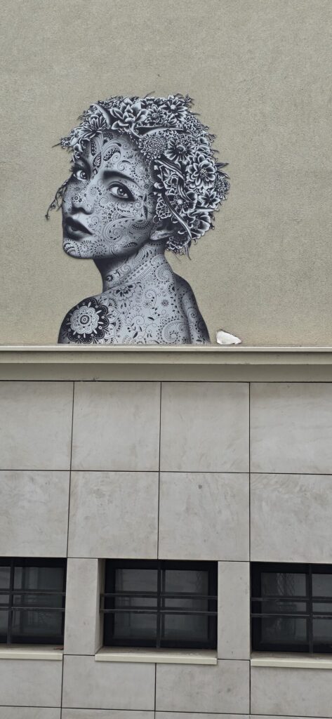 Close-up of a black and white ornamental paste-up on rue Gerard showing a woman with a crown of drawn flowers and paisley patterned skin, beside a tiled modern wall
