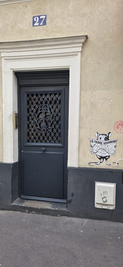 Doorway of number 27 rue des Cinq Diamants with a stencilled rat above the frame reading a map of Paris titled Le Paris Gourmand, and a green painted handprint on the junction box beside it