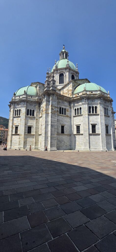 The pale stone facade and green domed cupola of Como Cathedral seen from below against a deep blue sky, with the dark cobbles of the piazza in the foreground