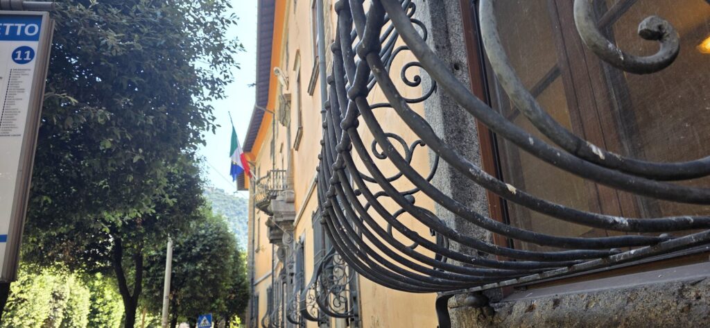 Close view of an ornate wrought iron balcony bracket, its scrolls curling out from the wall, with a street sign and trees out of focus behind