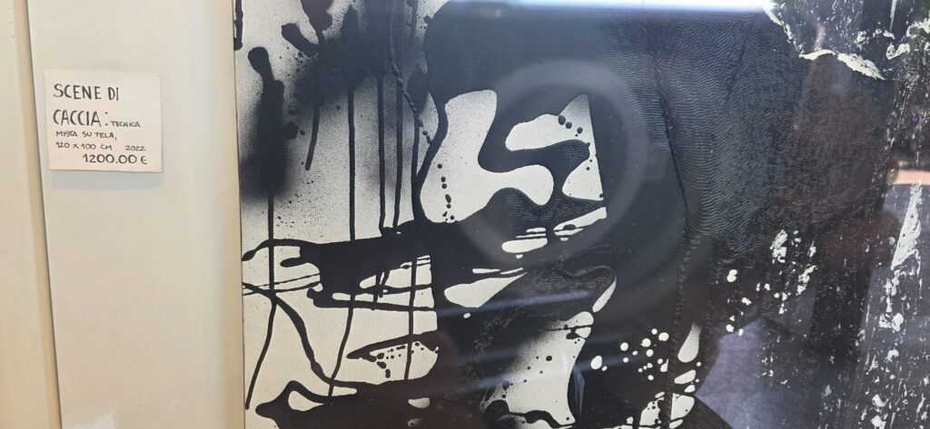 A large black and white abstract canvas behind glass in a gallery window, poured and dripped ink shapes on a white ground, with a small handwritten card beside it giving the title, technique, size, year and price