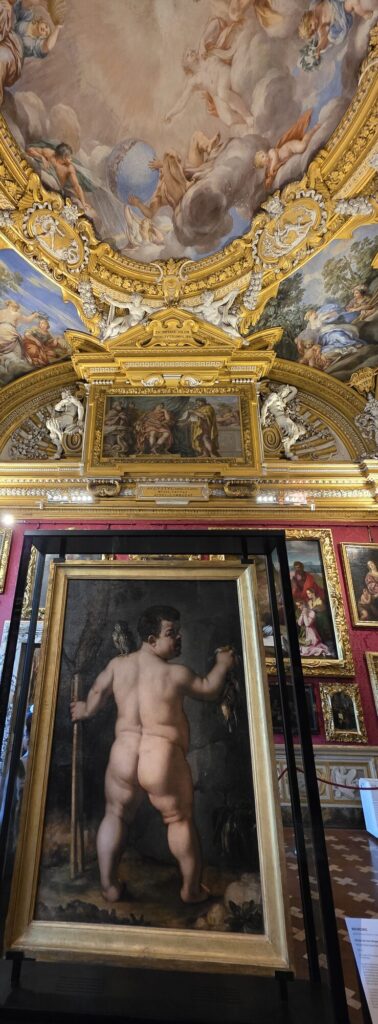 A large old painting standing free in a metal frame in the middle of a gallery, showing the back view of a naked man holding a staff and a dead bird, beneath a gilded and frescoed ceiling with red damask walls beyond