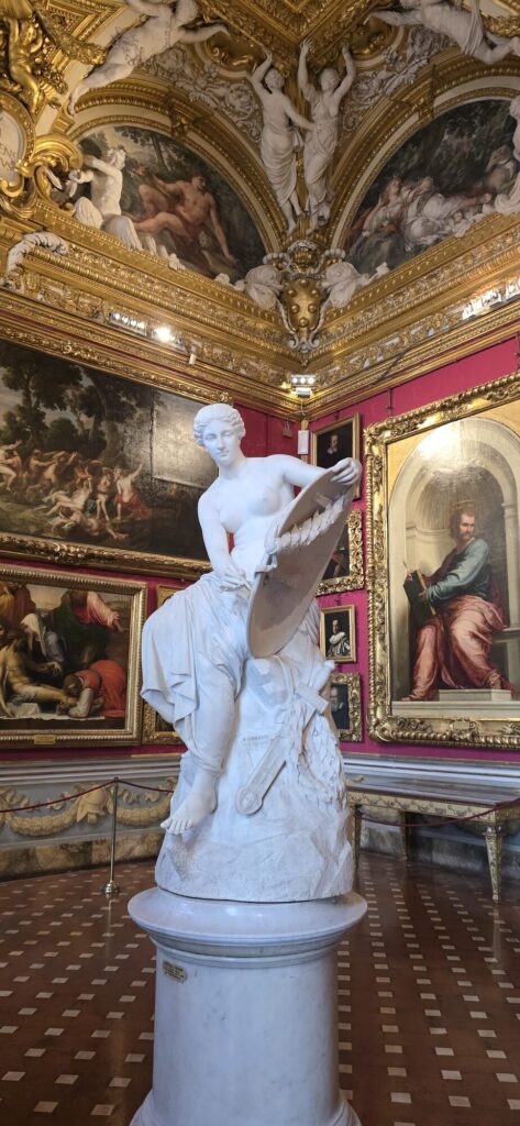 A white marble statue of a seated woman with a shield and drapery on a round pedestal, in a gallery with deep red walls, gilt framed paintings and an elaborate gilded and frescoed ceiling