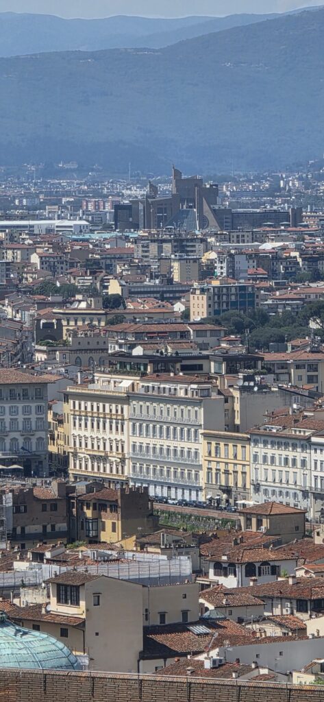 View across the terracotta rooftops of Florence from a height, with the tower and dome of the city rising above the tiles and blue hills behind