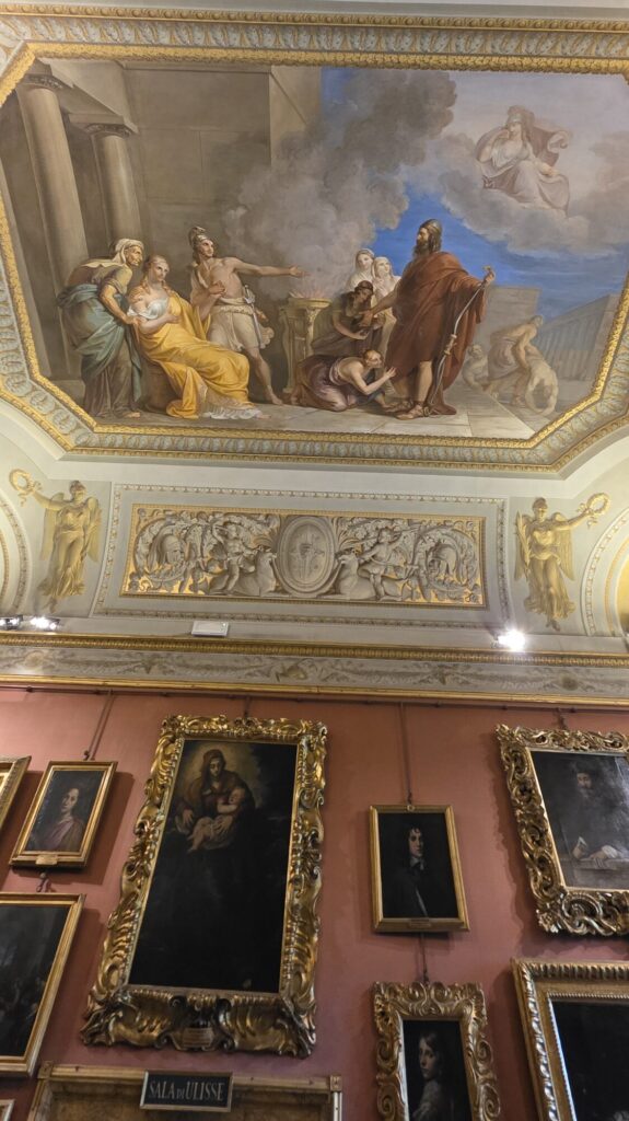 Gallery room with a painted ceiling showing a mythological scene, a grisaille frieze below it, and pink walls hung with many gilt framed portraits and religious paintings at different heights