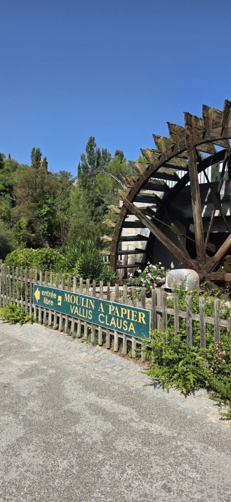 The huge wooden paddle wheel of the Vallis Clausa paper mill behind a chestnut paling fence, with a green sign reading Moulin a Papier Vallis Clausa and entree libre