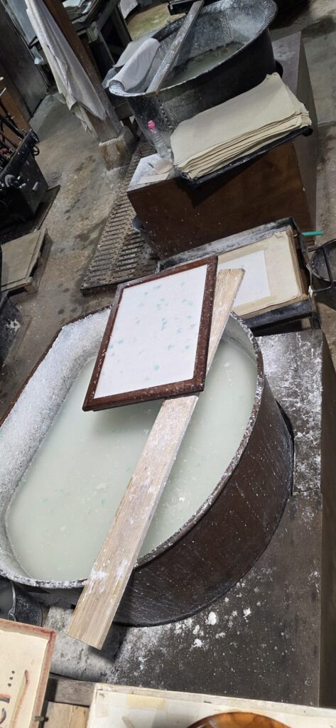 A vat of pale paper pulp at the Vallis Clausa mill with a wooden mould and deckle resting across it holding a freshly formed sheet with green petals set into it, and stacks of pressed sheets on the bench behind